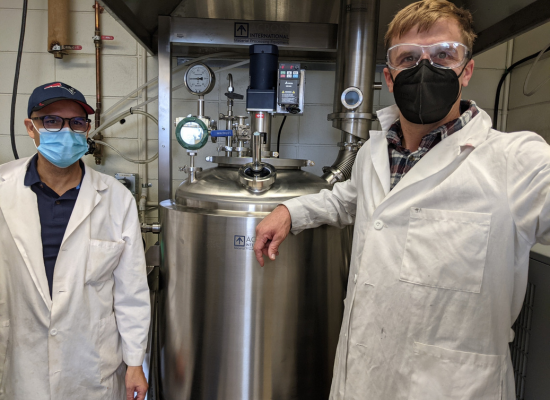 Two men in lab coats and face mask in front of a metal, cylindric bio-reactor